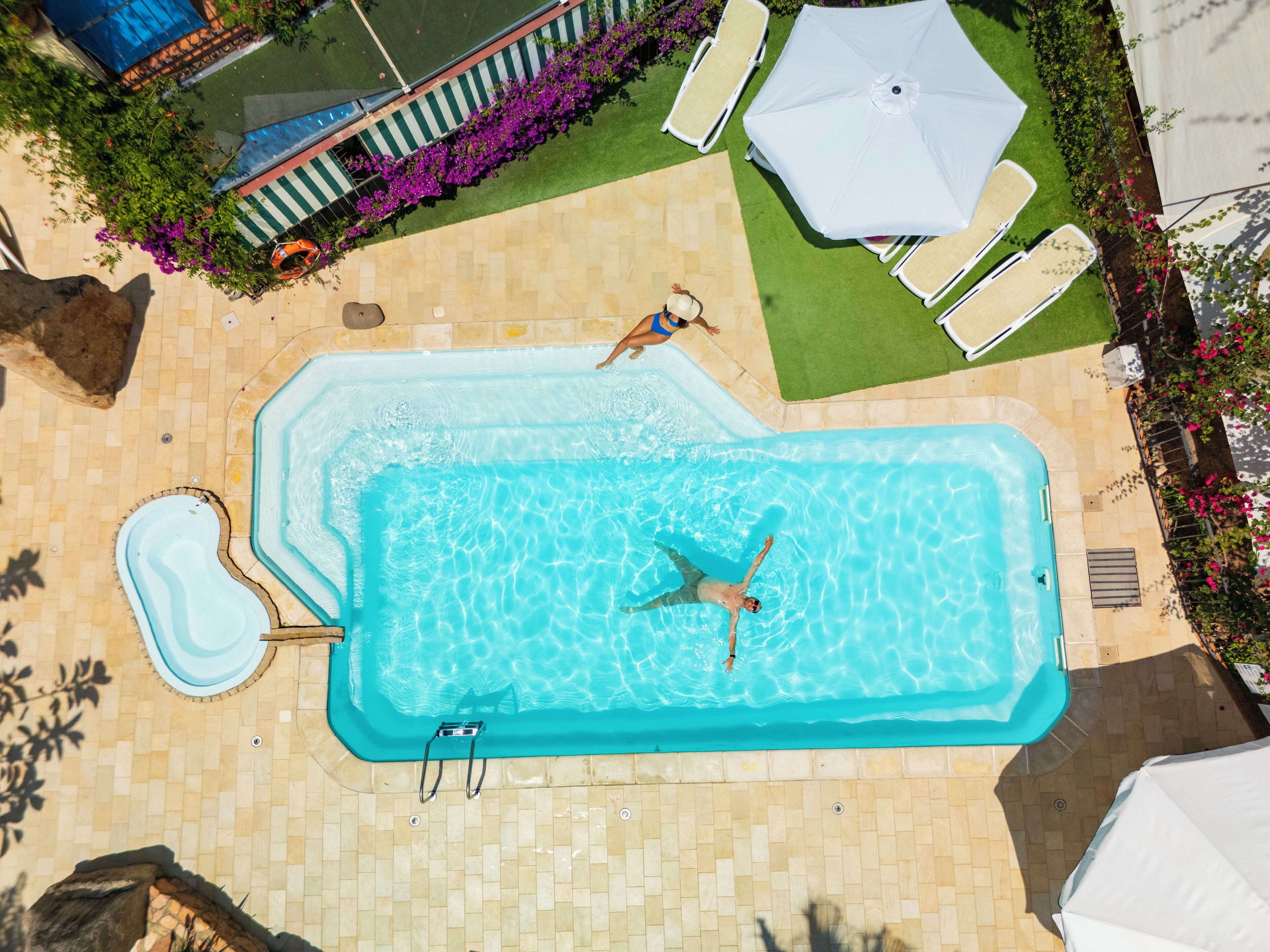 A rectangular swimming pool with a diving board, surrounded by a tiled deck and a small garden area with flowers and a lawn.
