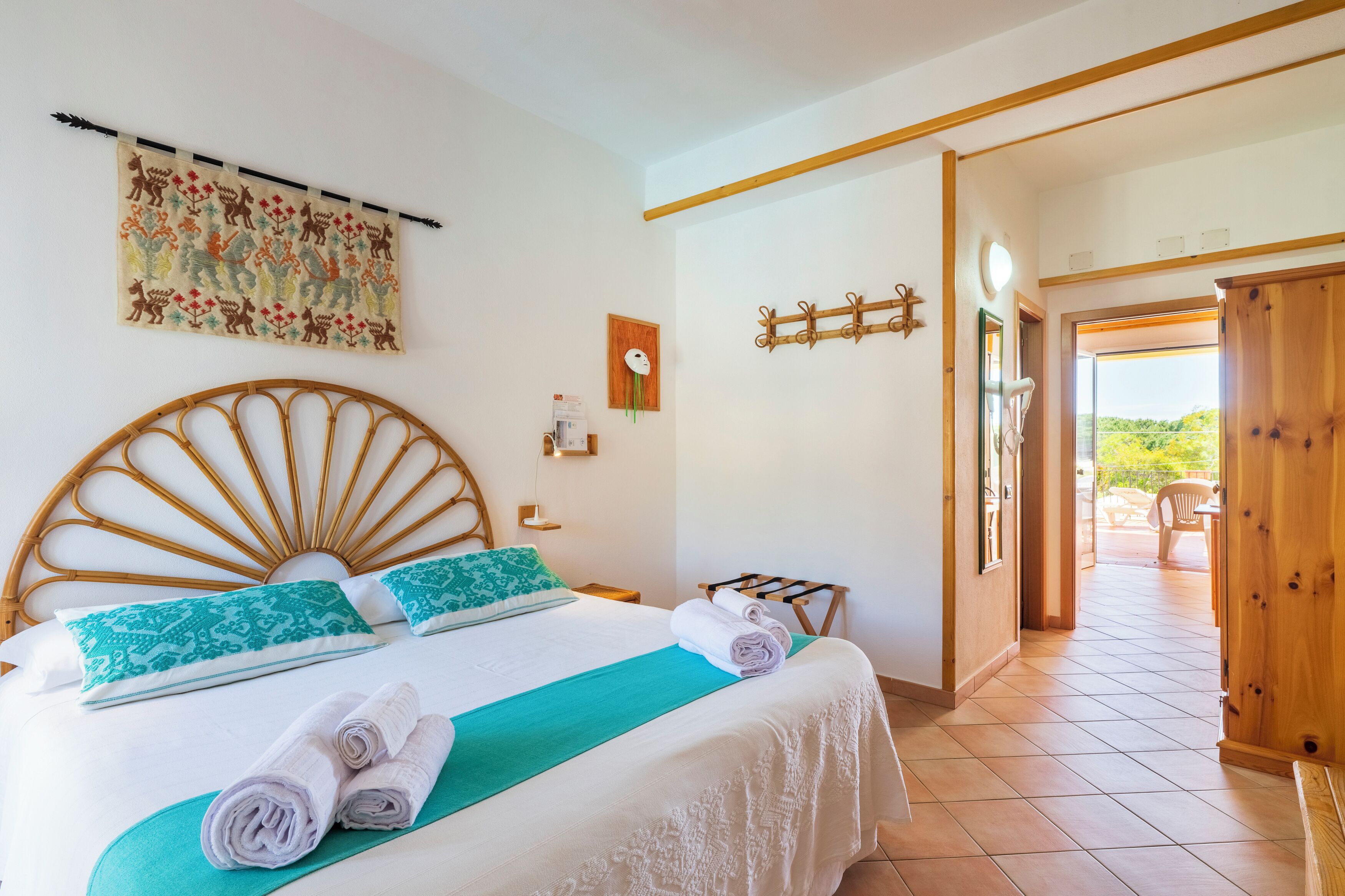 A neatly made bed with a headboard, a wall-mounted artwork, a wooden coat rack, and a view of the outside through an open door.
