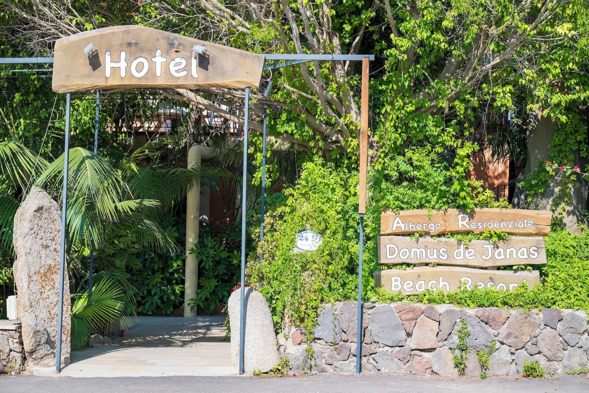 Ein hölzernes Schild mit der Aufschrift 'Hotel', eine Steinmauer und ein Schild, das 'Albergo Residenziale Domus de Janus Beach Resort' anzeigt.