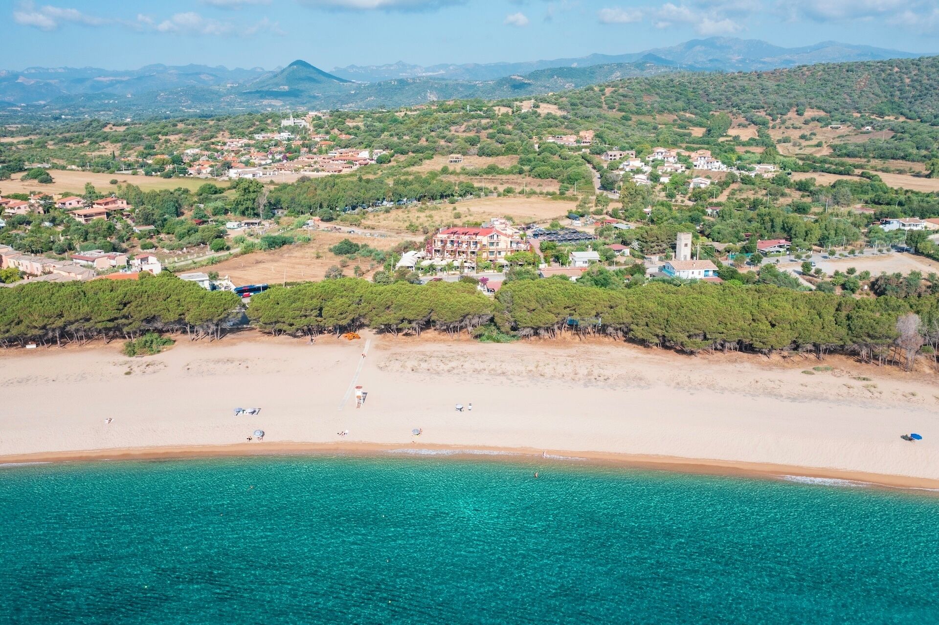 An aerial view of a coastal town with a prominent building surrounded by greenery, a sandy beach, and clear turquoise waters.