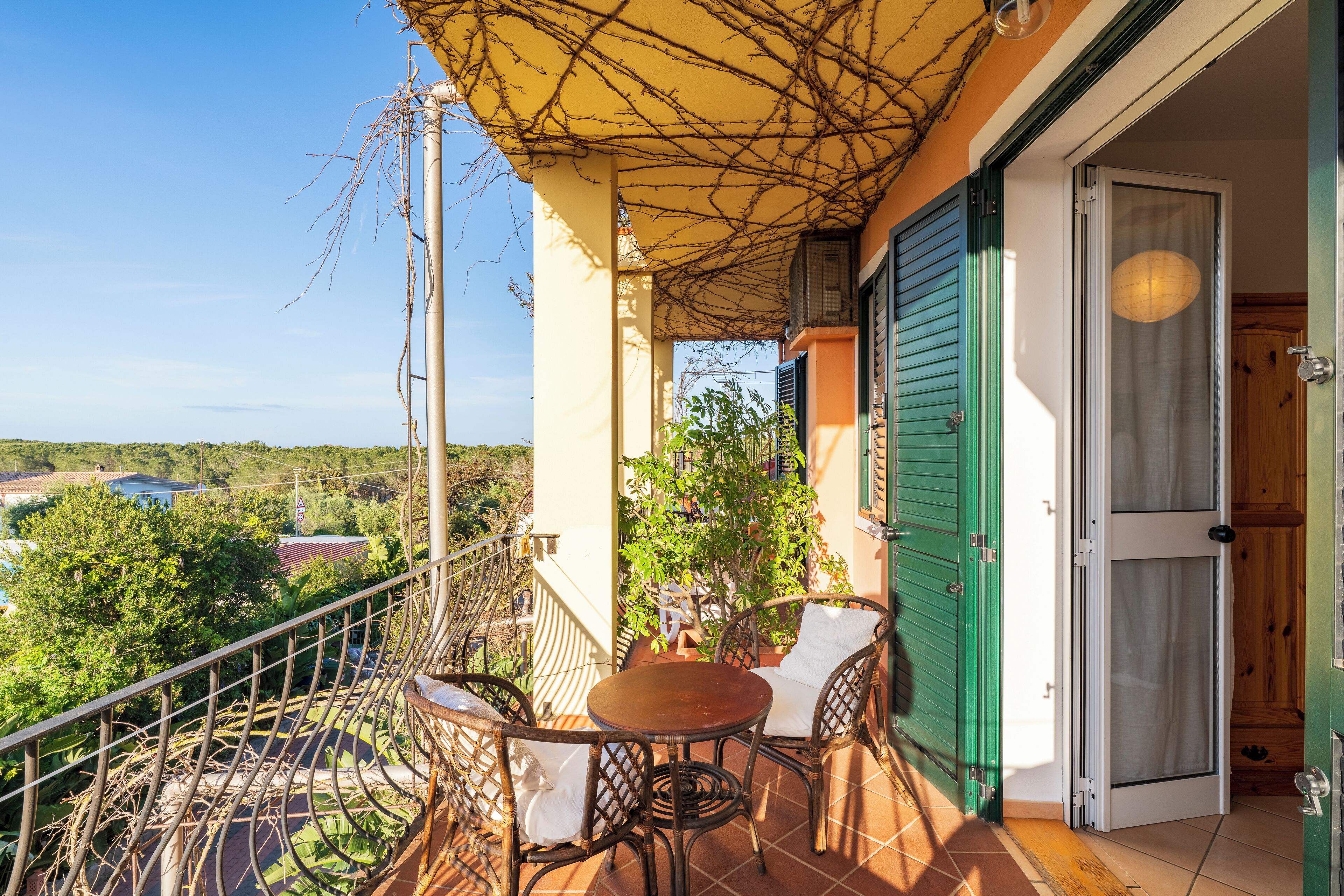 A balcony with a table and chairs, a view of the surrounding area, and a door with a green shutter.