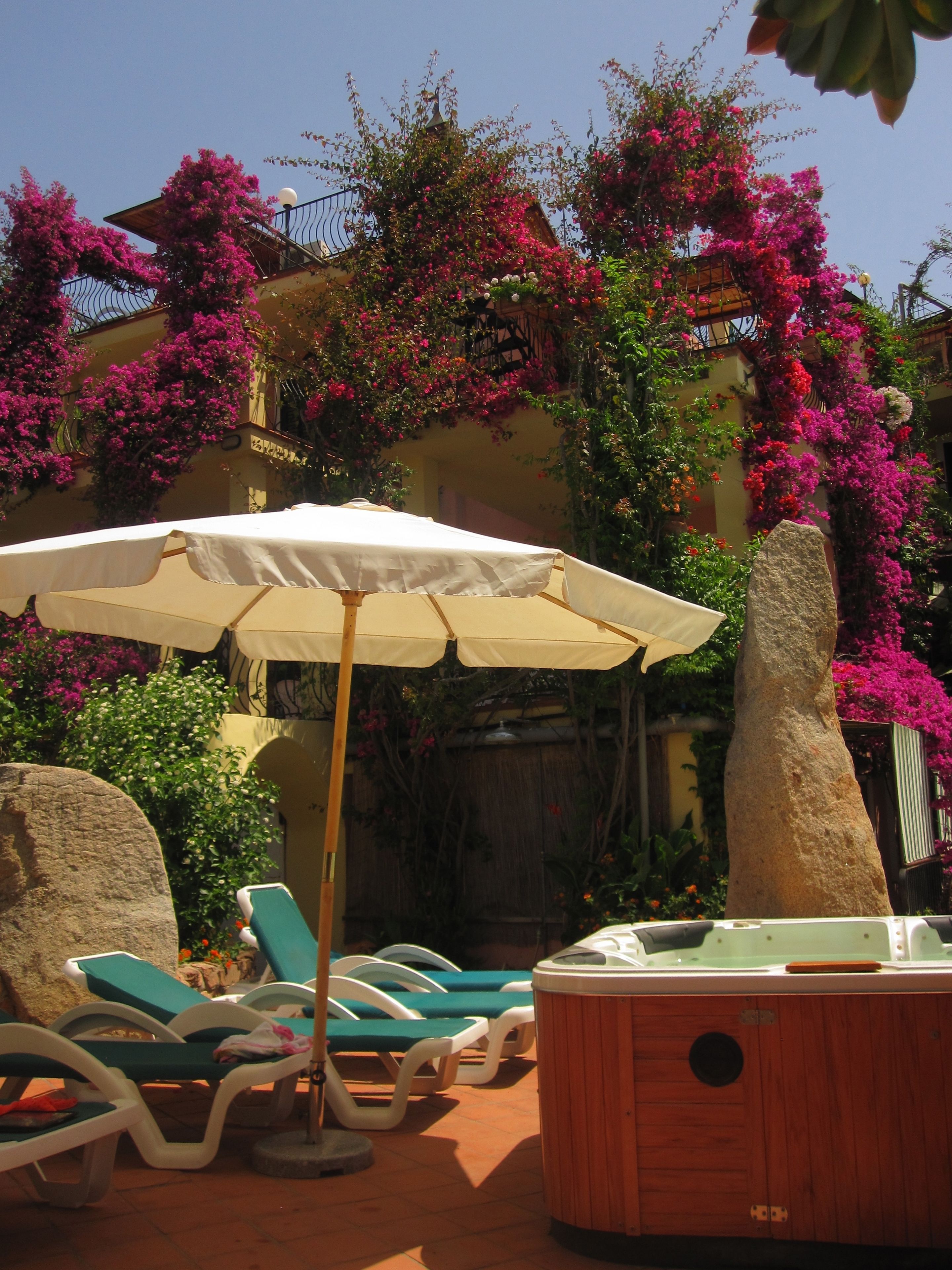 A pool area with lounge chairs and a hot tub, surrounded by vibrant bougainvillea flowers and a stone pillar.