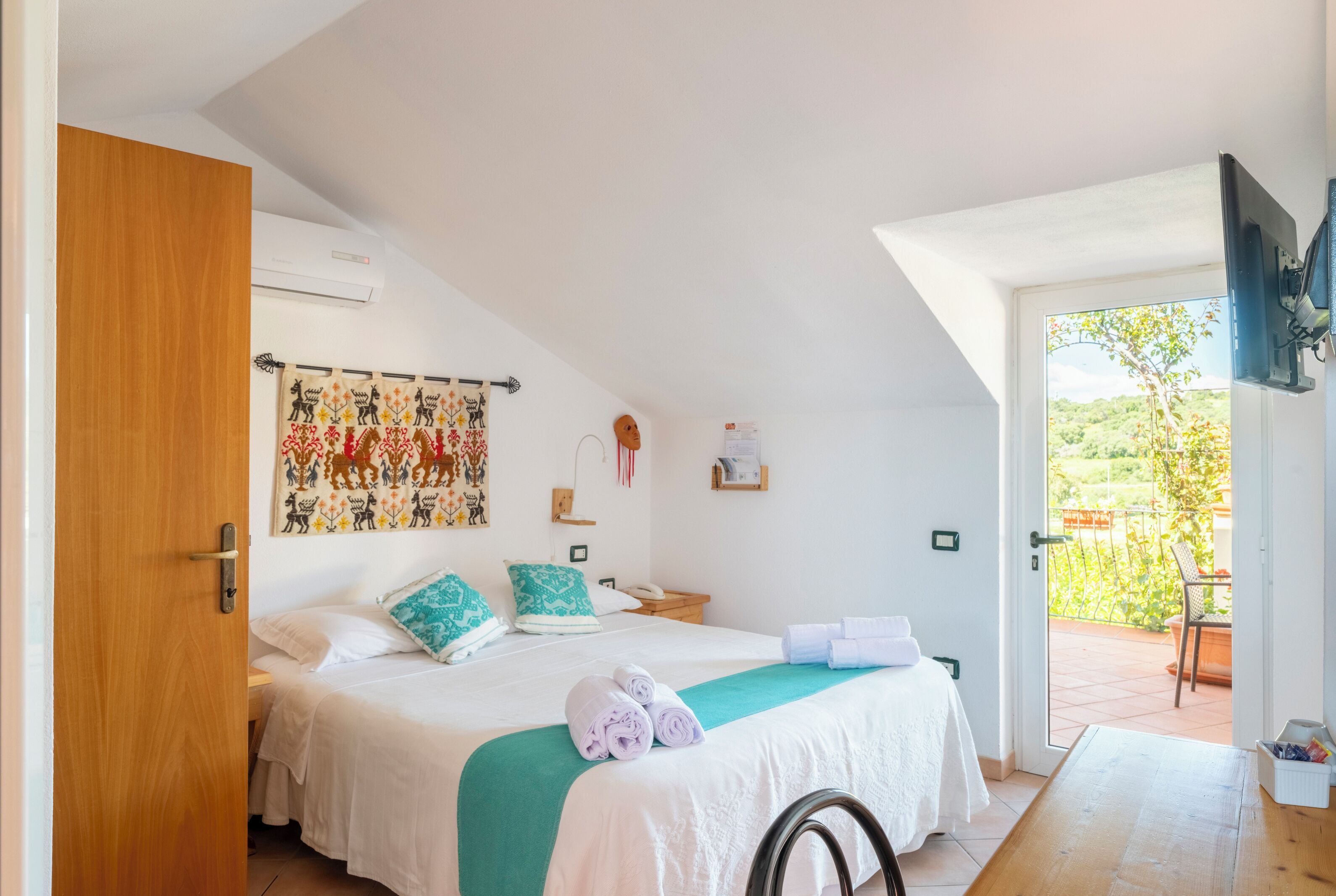 Un dormitorio con una cama, una puerta de madera, un ventilador de techo, una ventana y vistas a la vegetación exterior.