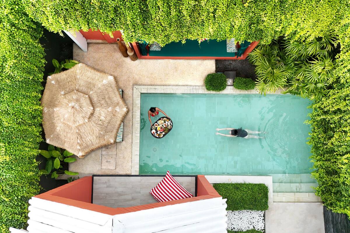 Vista aérea de una piscina rodeada de abundante vegetación, con una persona nadando y otra en una plataforma flotante.