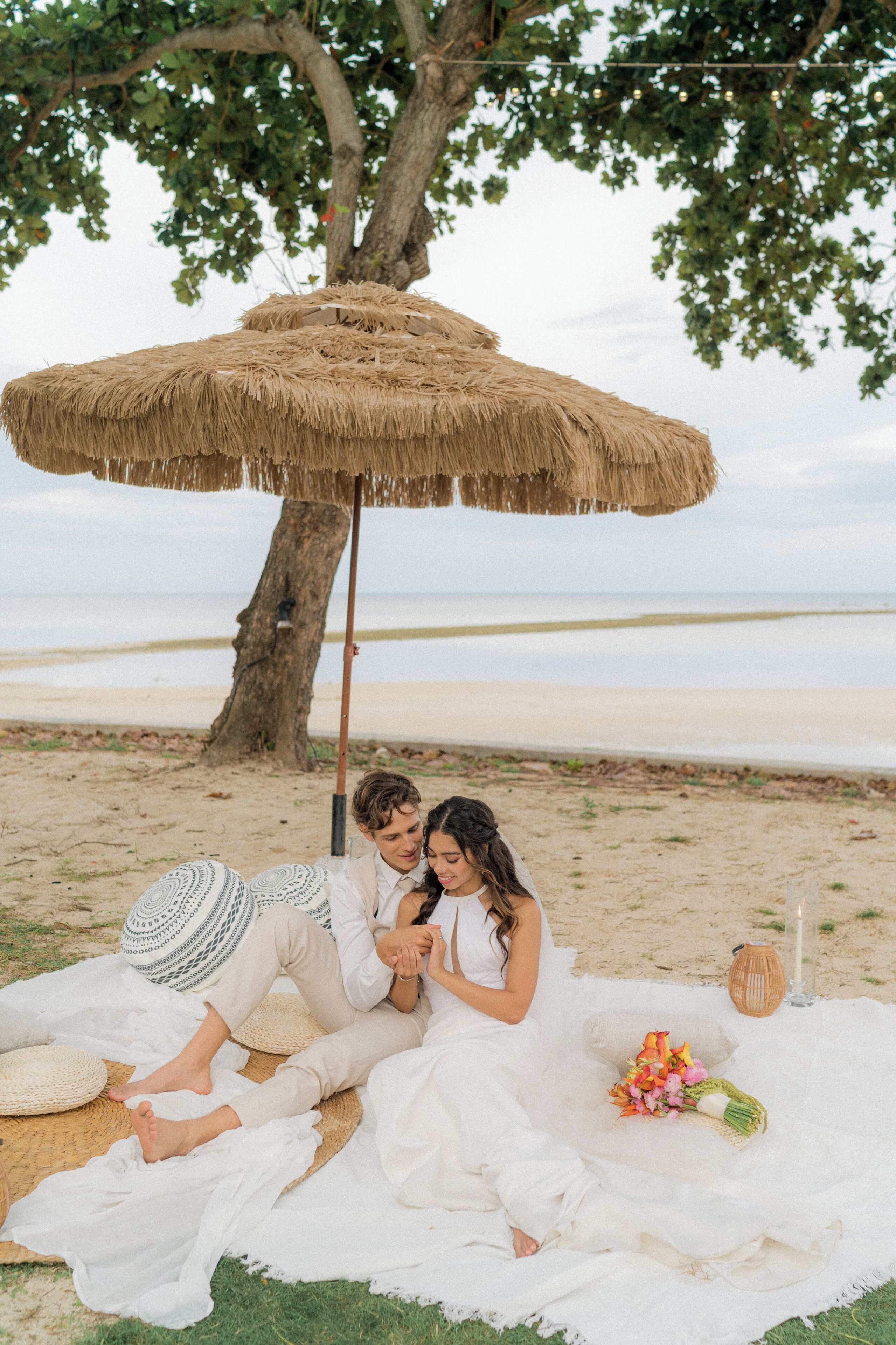Escena de playa con una sombrilla de paja, una manta y un ramo de flores.