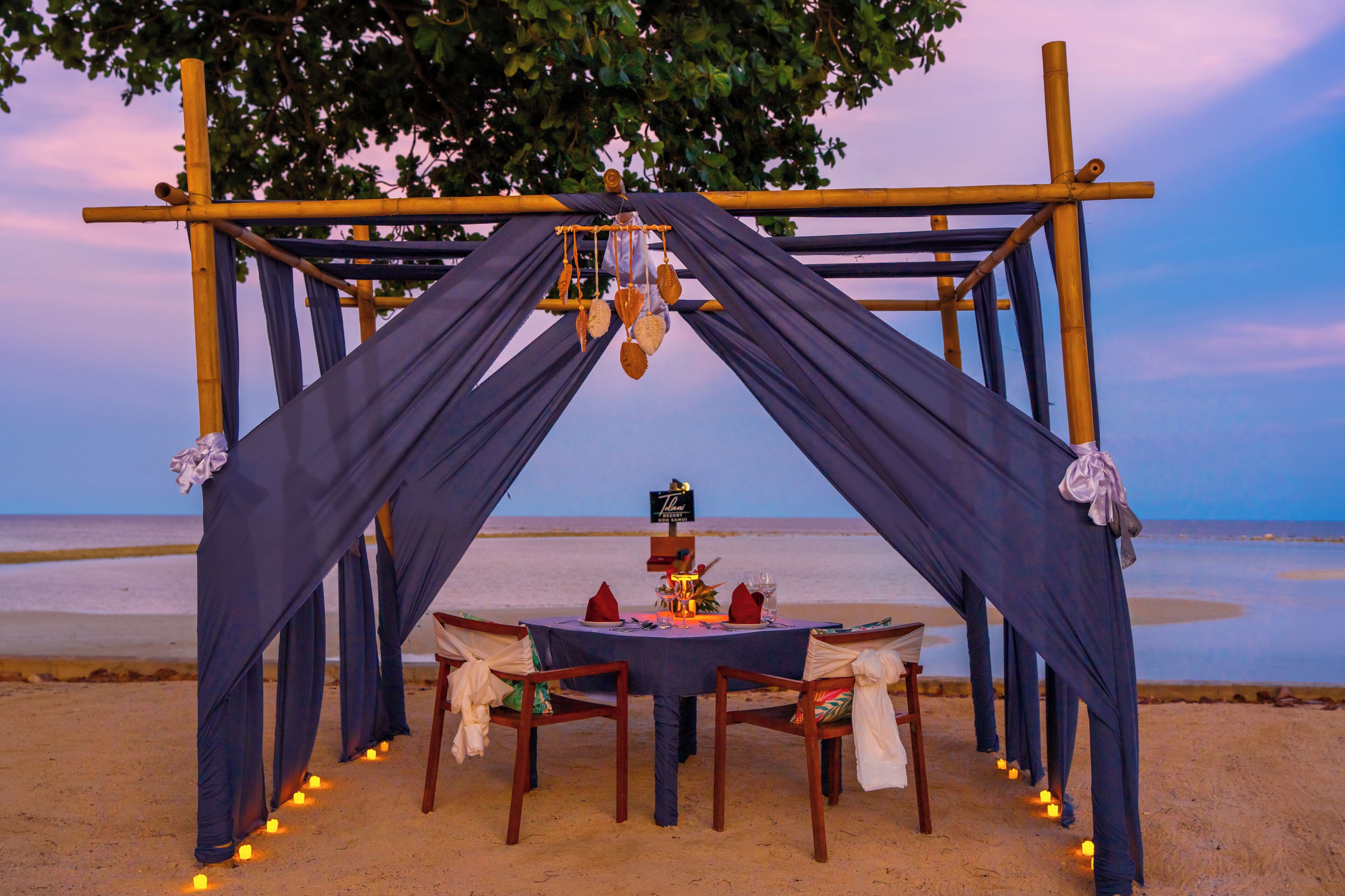Montaje para comer en la playa con una mesa y sillas bajo un arco de bambú, adornado con velas y un letrero.