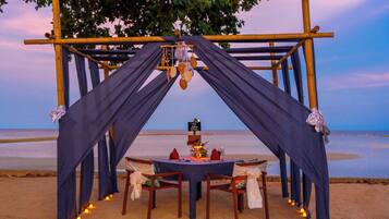Montaje para comer en la playa con una mesa y sillas bajo un arco de bambú, adornado con velas y un letrero.