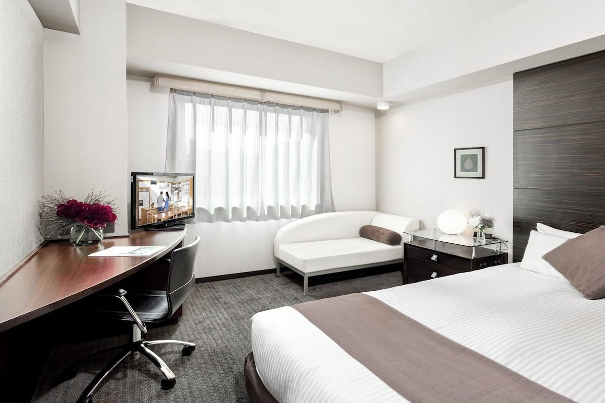 A hotel room with a bed, a desk, a chair, a TV, and a window with curtains.