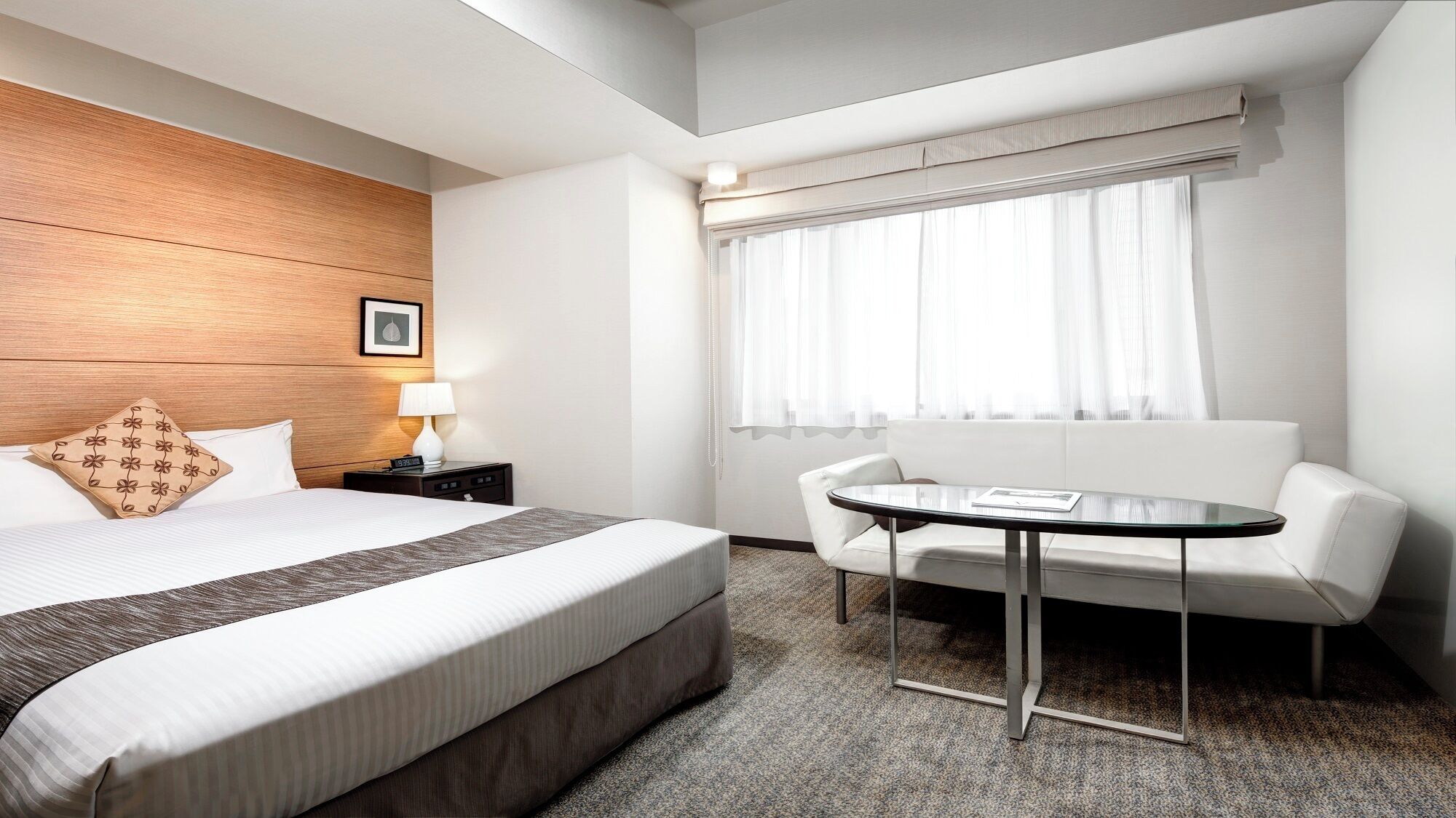 A hotel room with a bed, bedside table, lamp, and a seating area with a table and chair.