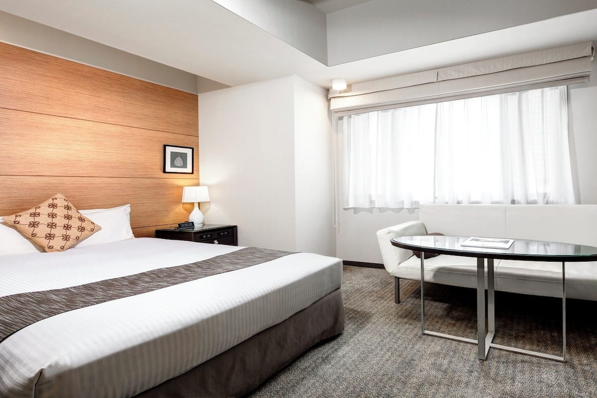 A hotel room with a bed, bedside table, lamp, and a seating area with a table and chair.