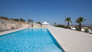 A swimming pool with clear blue water, surrounded by a tiled deck, a white tent, and palm trees. - Villa Principe di Belmonte (Ispica)