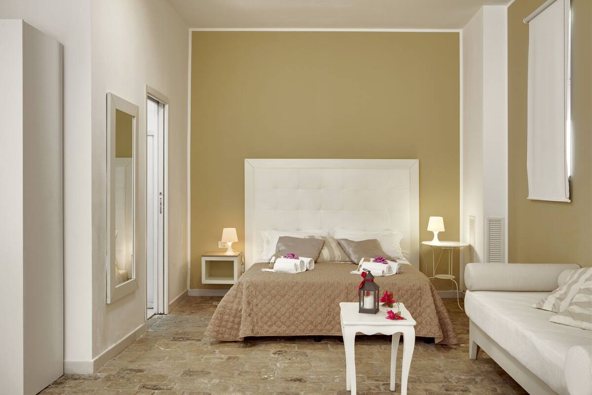 A hotel room with a large bed, two bedside tables with lamps, a white sofa, and a small table with a lantern and flowers.