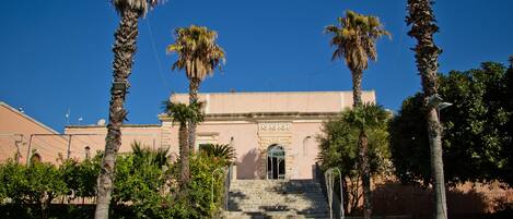 A building with a central staircase leading to a doorway, surrounded by palm trees and greenery.