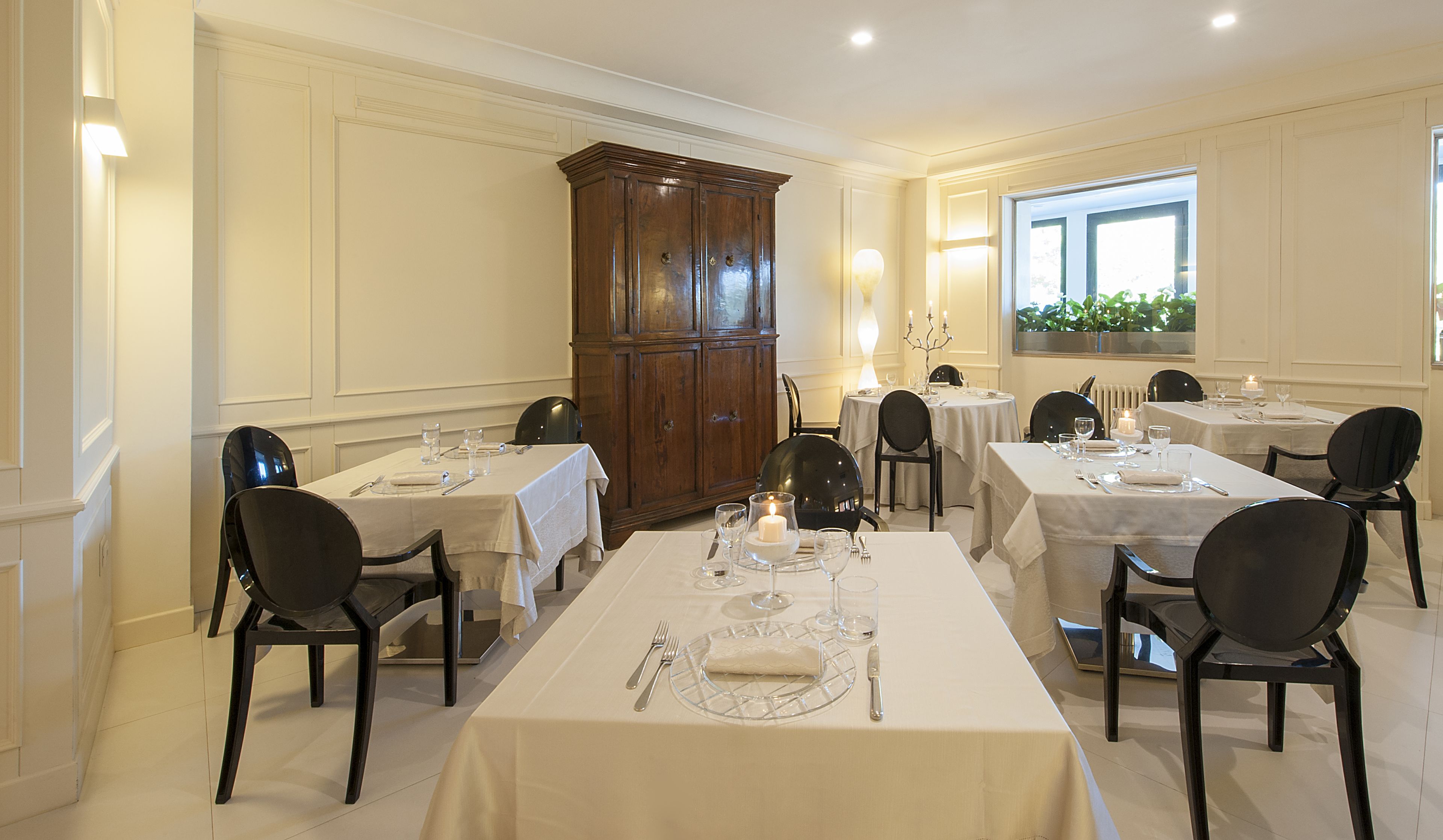 A formal dining room with multiple tables set for service, a large wooden cabinet, and a window with a view of greenery.