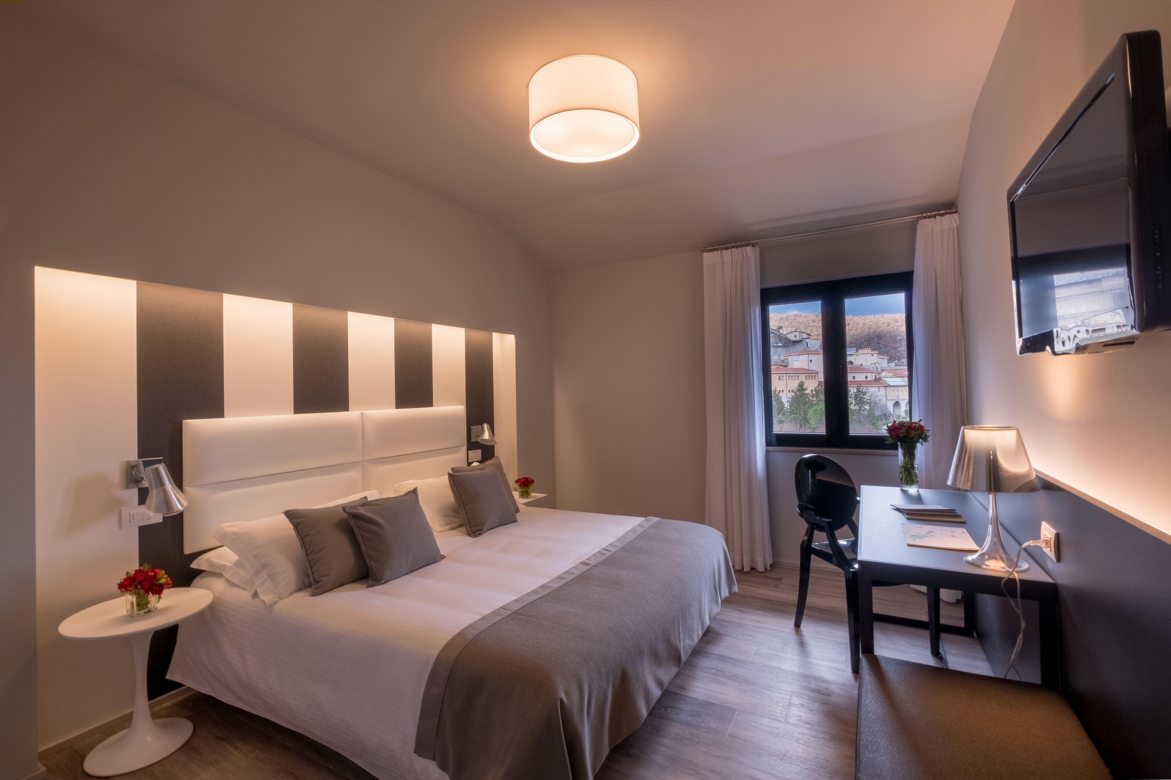 Quarto de hotel moderno com uma cama grande, mesas de cabeceira, escrivaninha e televisão.