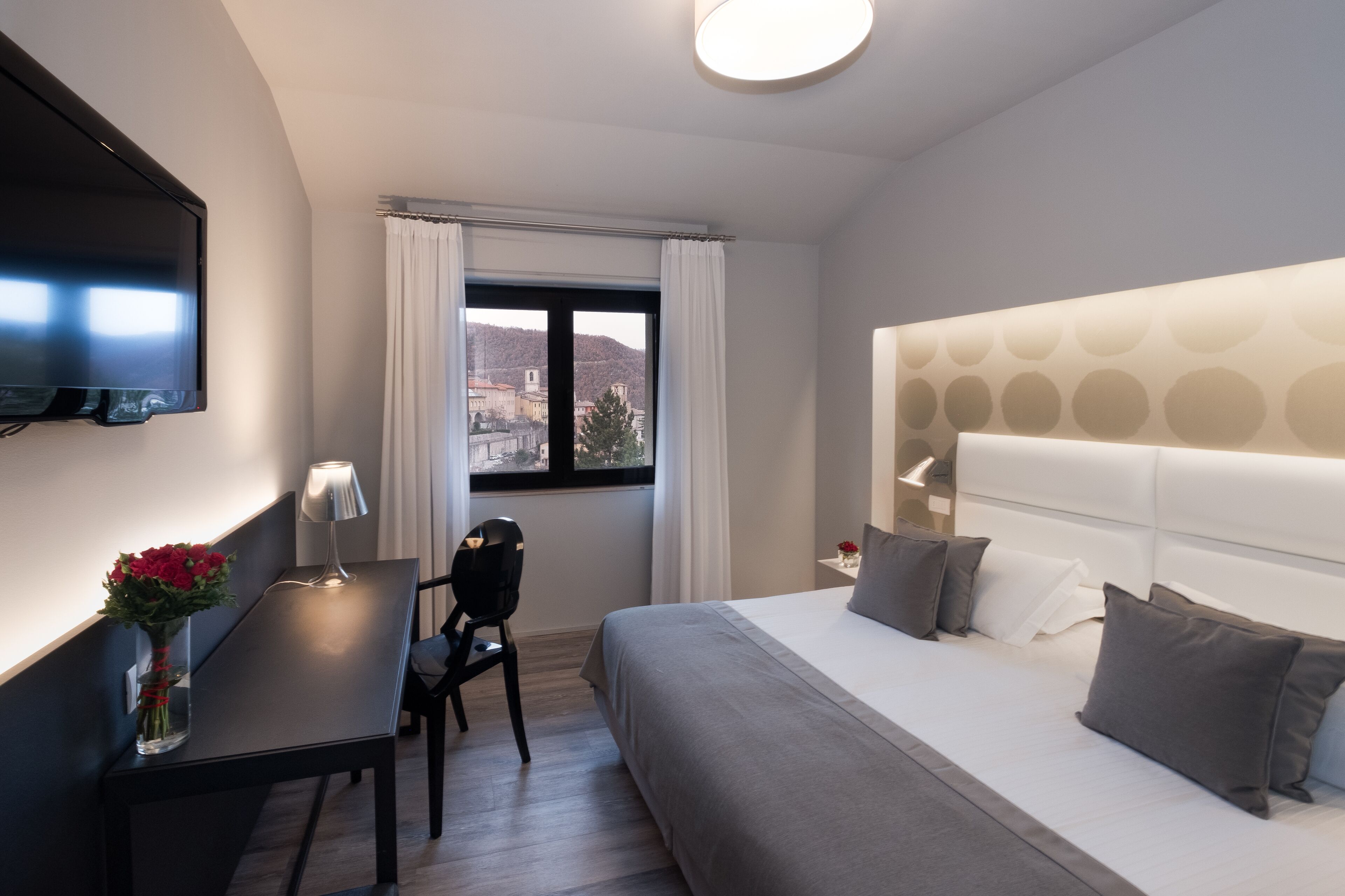 A modern hotel room with a bed, a desk, a chair, a TV, and a window with curtains.