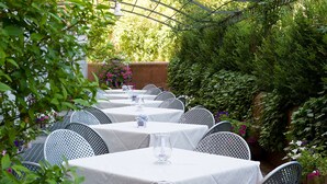 A row of tables with white tablecloths and metal chairs, arranged outdoors with greenery and flowers around. - Hotel Monte Meraviglia (Cascia)