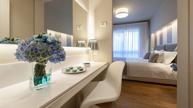 A modern hotel room with a large bed, a bedside table with a vase of flowers, a desk with a lamp, and a large mirror. - Hotel Monte Meraviglia (Cascia)