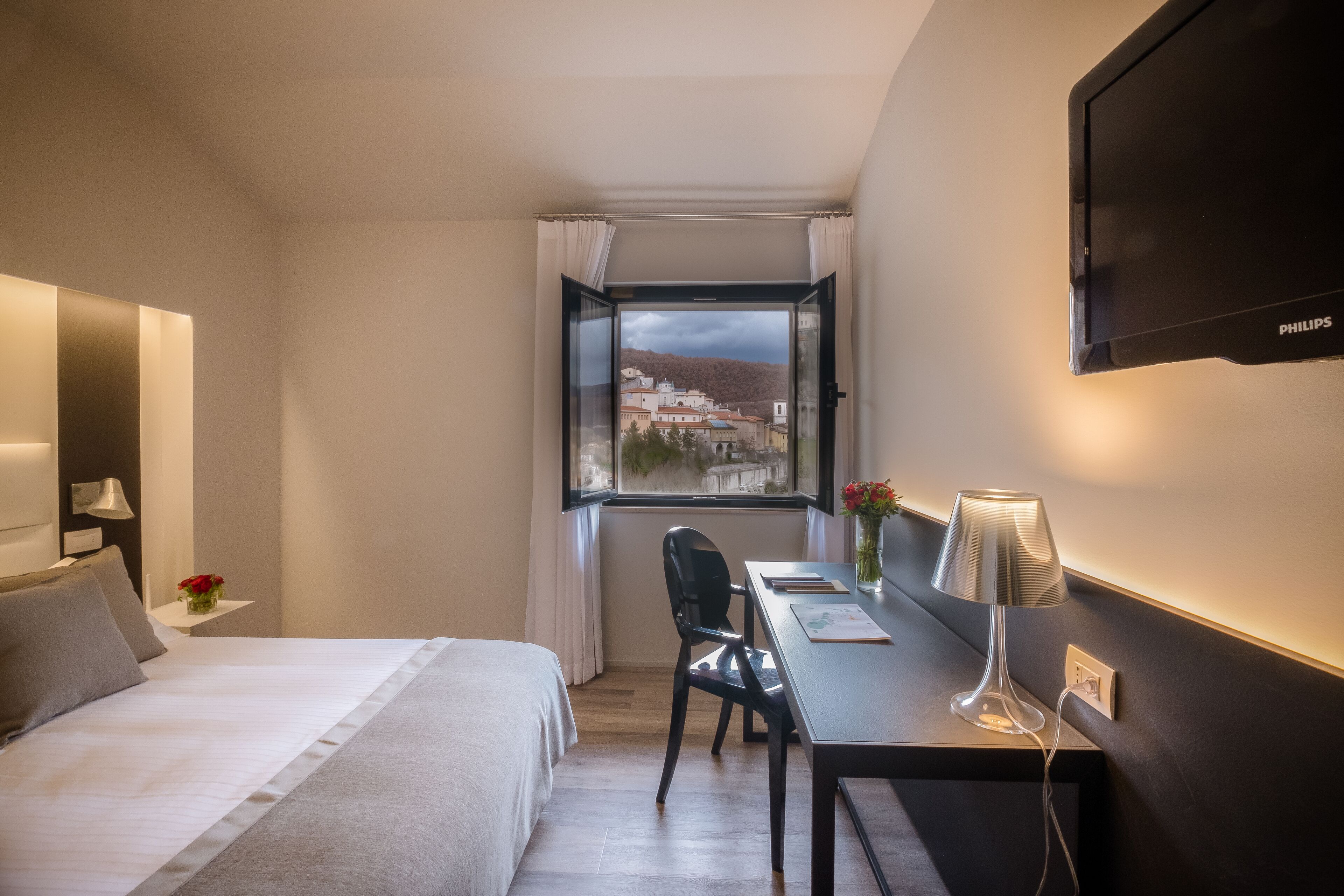 Et hotelværelse med en seng, et skrivebord med lampe, en stol og et fjernsyn ophængt på væggen.