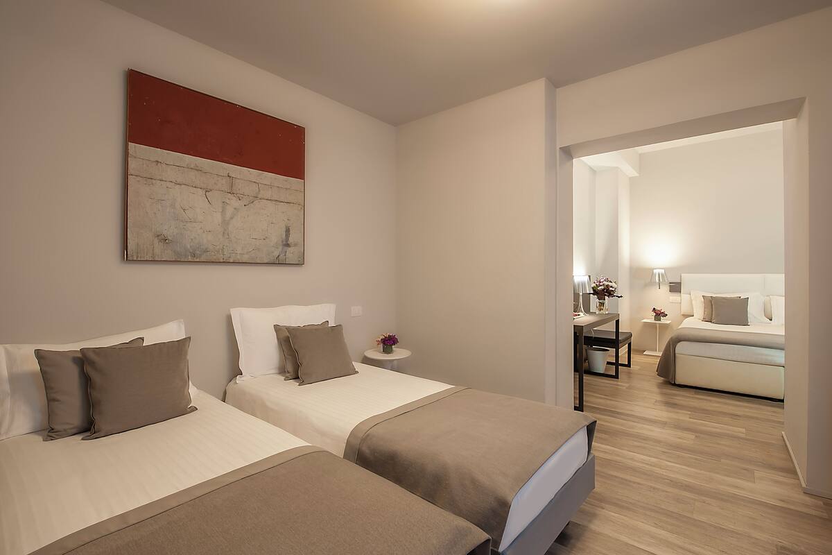 Ein Hotelzimmer mit zwei Betten, einem Schreibtisch und einem Stuhl. Auf einem kleinen Tisch steht eine Blumenvase.