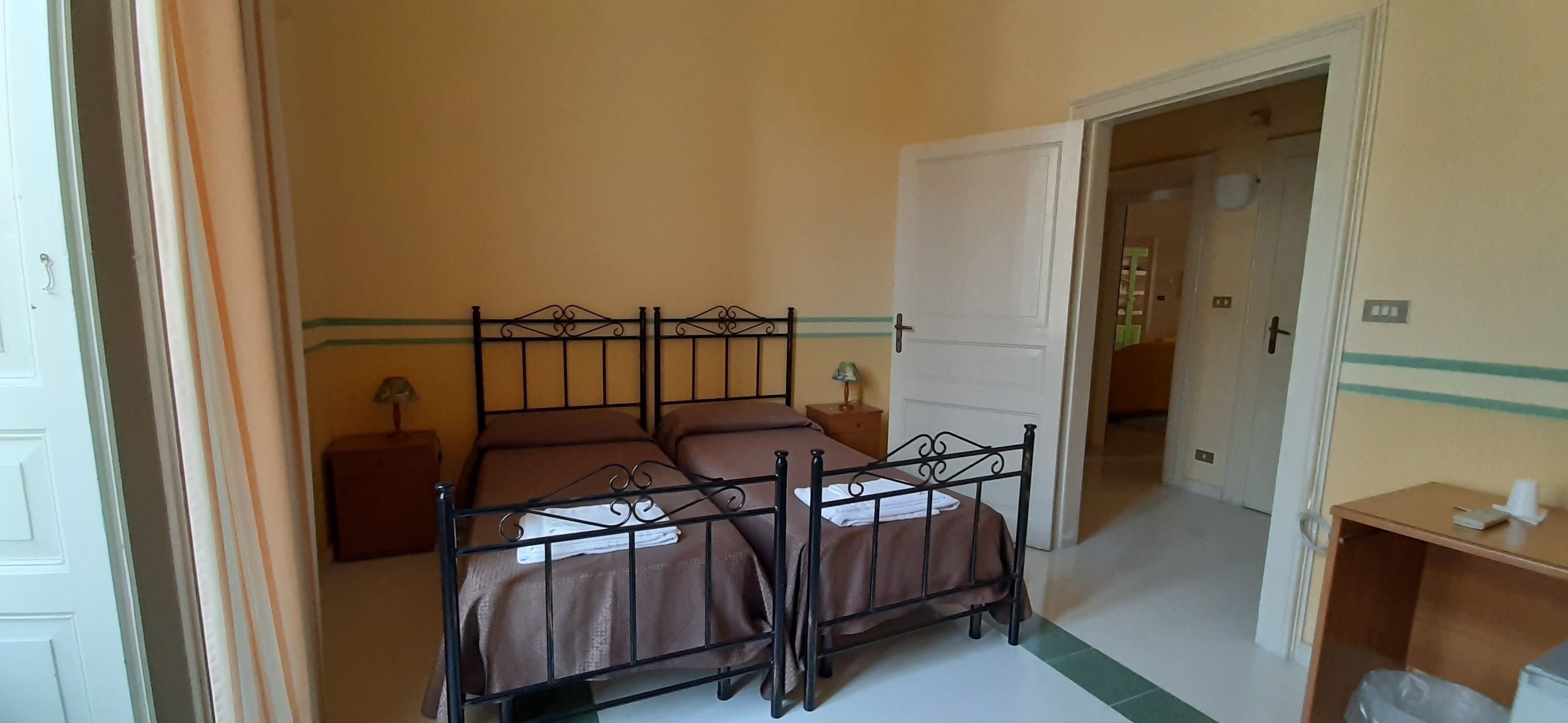 Un dormitorio con dos camas individuales, una mesita de noche, una lámpara y una puerta que da a otra habitación.