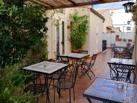 Um pátio externo com piso de azulejos, mesas e cadeiras de metal e plantas em vasos.