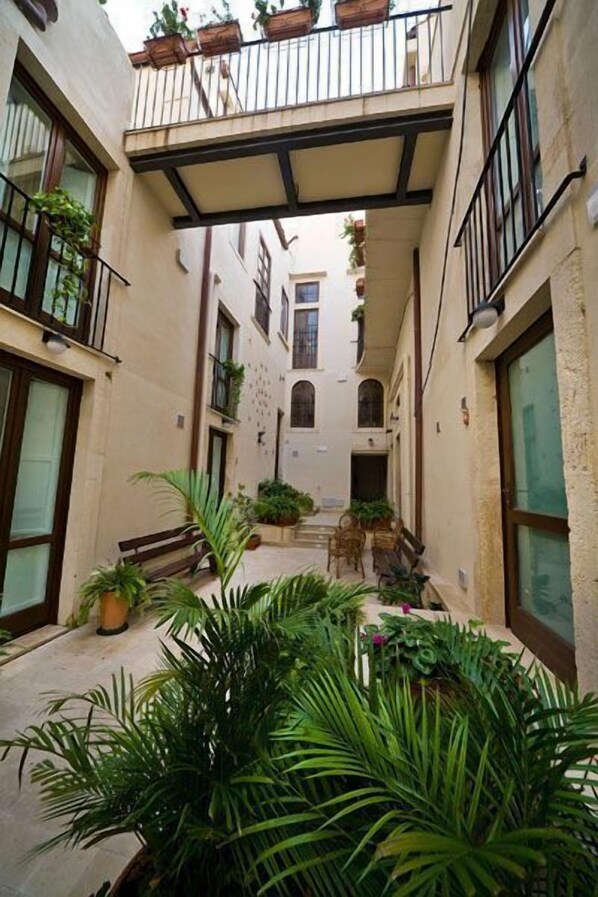 A narrow courtyard with potted plants and benches, surrounded by buildings with balconies and large windows.