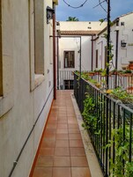 A narrow corridor with tiled flooring, a metal fence on one side, and a white building with a tiled roof in the background.