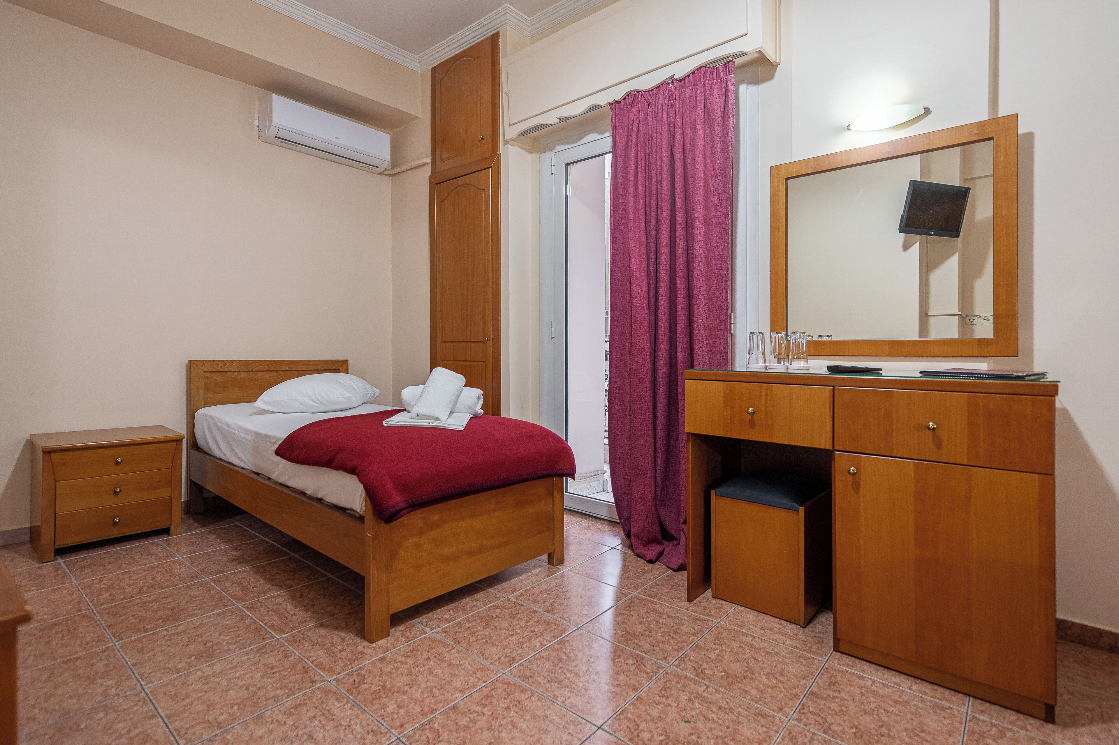 Une chambre avec un lit en bois, une table de chevet, une commode en bois avec un miroir et une porte-fenêtre coulissante avec un rideau violet.