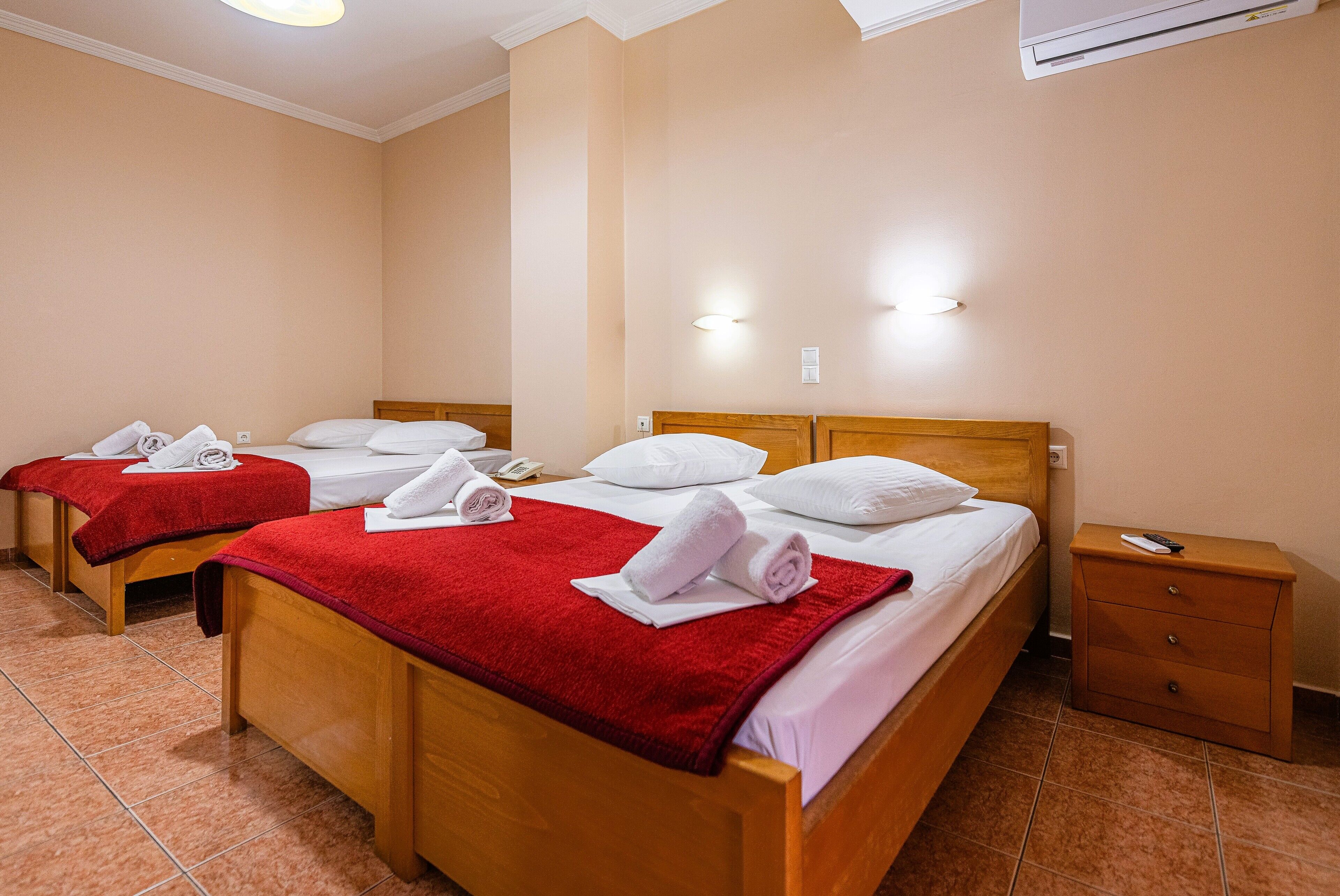A hotel room with three wooden beds, each with a red blanket and white towels. There is a wooden nightstand with a remote control and a drawer.