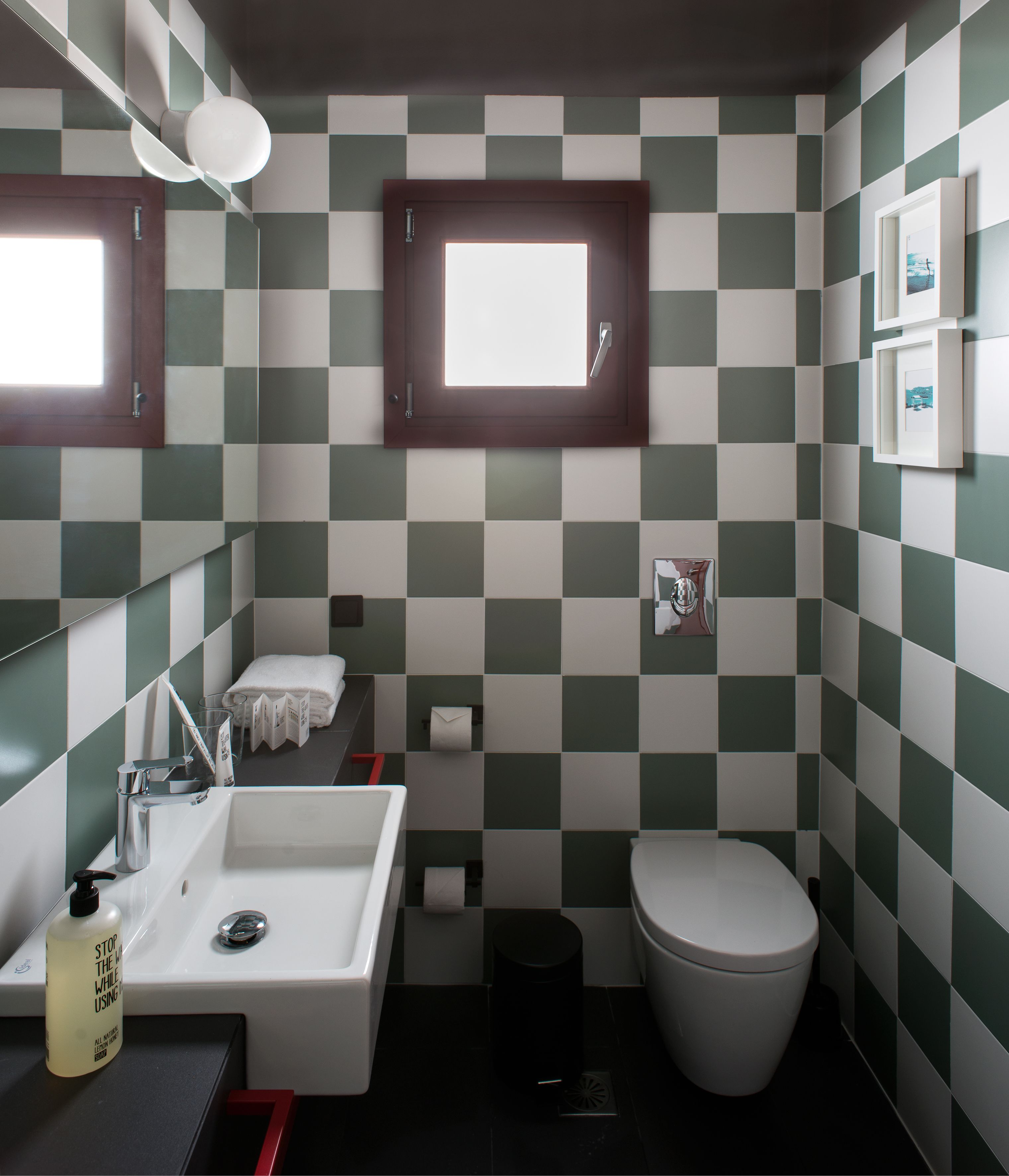 A bathroom with a checkered tile wall, a sink with a mirror, a toilet, and a window above the mirror.