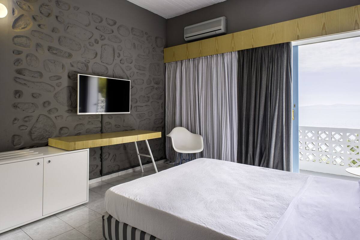 Une chambre d’hôtel moderne avec un mur au motif de pierres, un téléviseur à écran plat, un bureau en bois, une chaise blanche, un lit avec une couette blanche et une vue sur la mer par de grandes fenêtres.