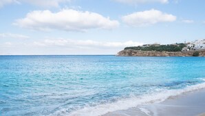 A beach with clear blue water, a sandy shore, and a coastal town in the background.