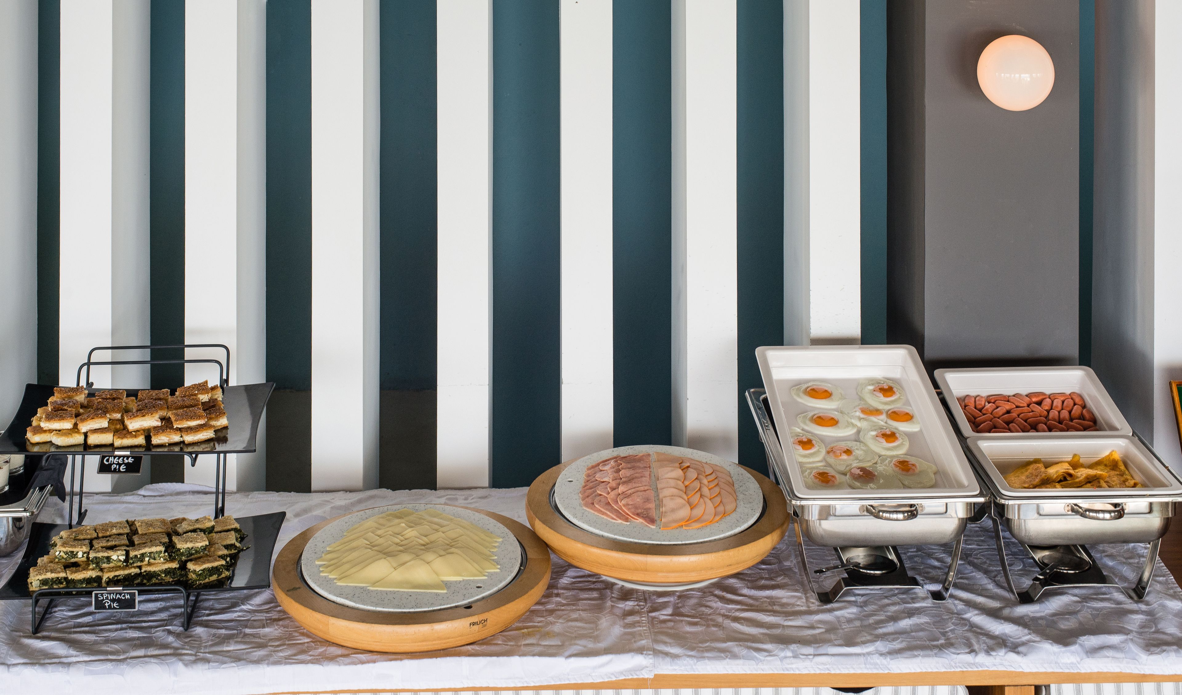 Una mesa de bufé con diferentes platillos, incluyendo empanadas de queso, espinacas y otras preparaciones, frente a un fondo de pared a rayas.