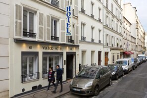 Um hotel com uma placa indicando "HOTEL VIATOR" e o número "61" na fachada. Há carros estacionados na rua em frente ao hotel.