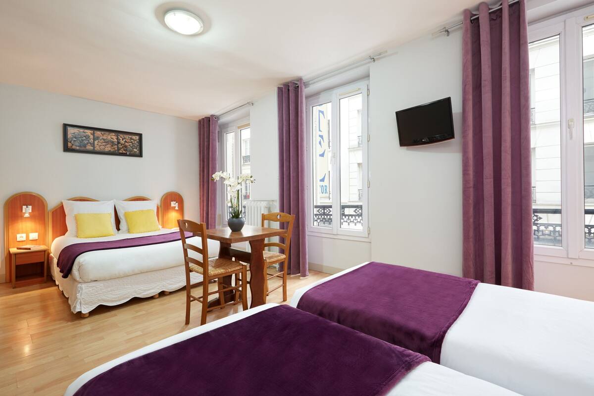 A hotel room with two beds, a dining table, chairs, a TV, and a window with curtains.