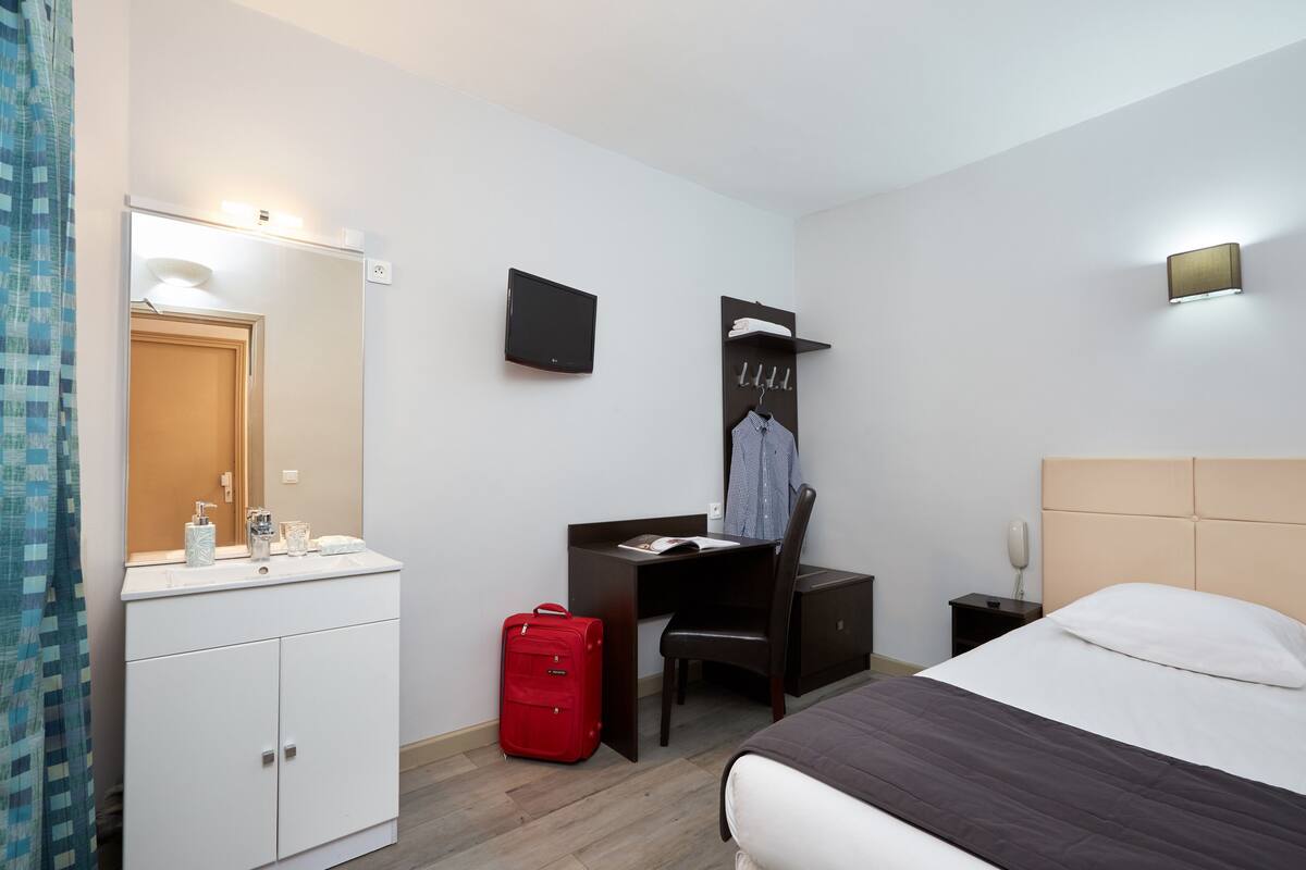 A hotel room with a bed, a desk with a chair, a TV mounted on the wall, a wardrobe, a red suitcase, and a bathroom with a mirror and sink.