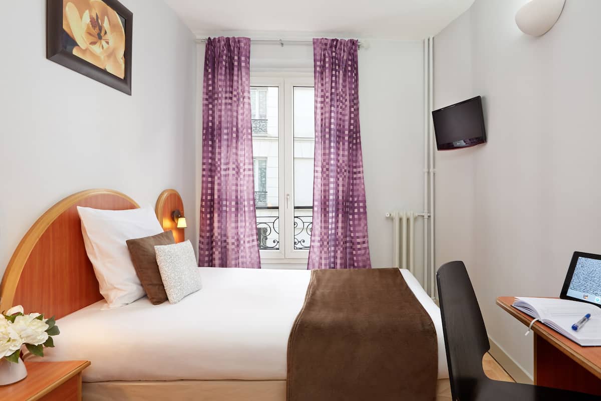 A hotel room with a bed, a desk, a chair, a TV, and a window with curtains.
