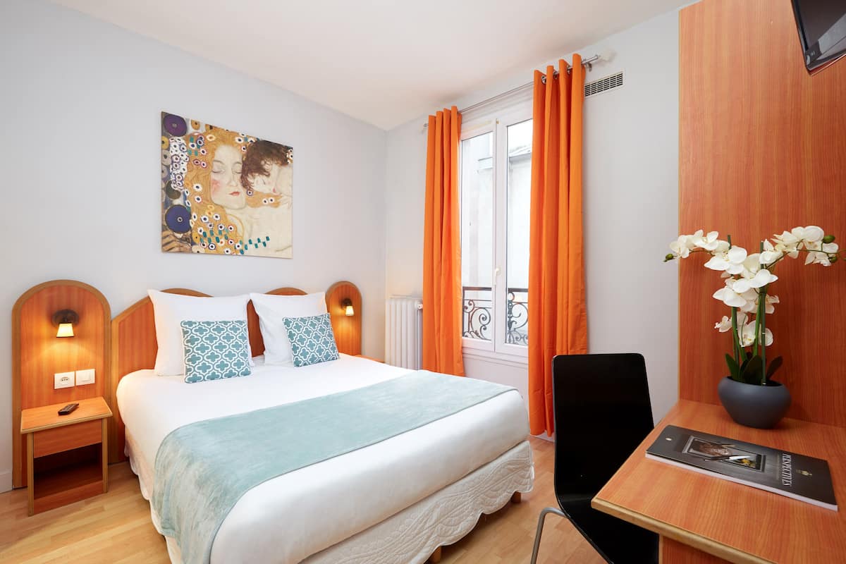A hotel room with a bed, a desk, a chair, a vase with flowers, a painting on the wall, and orange curtains.