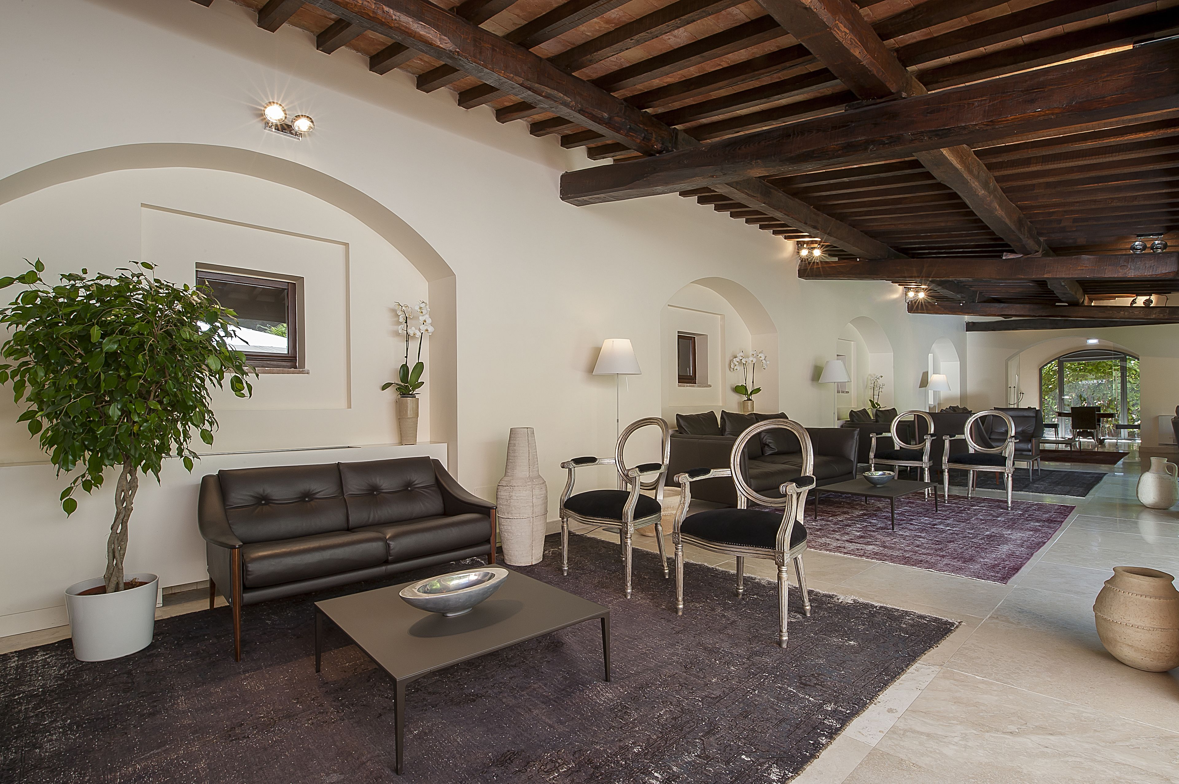 Amplia sala de estar con un sofá de cuero oscuro, una mesa de centro y una gran planta en maceta. Hay varias opciones de asiento, incluyendo un sofá y sillas. La habitación cuenta con vigas de madera expuestas y un ventanal grande.