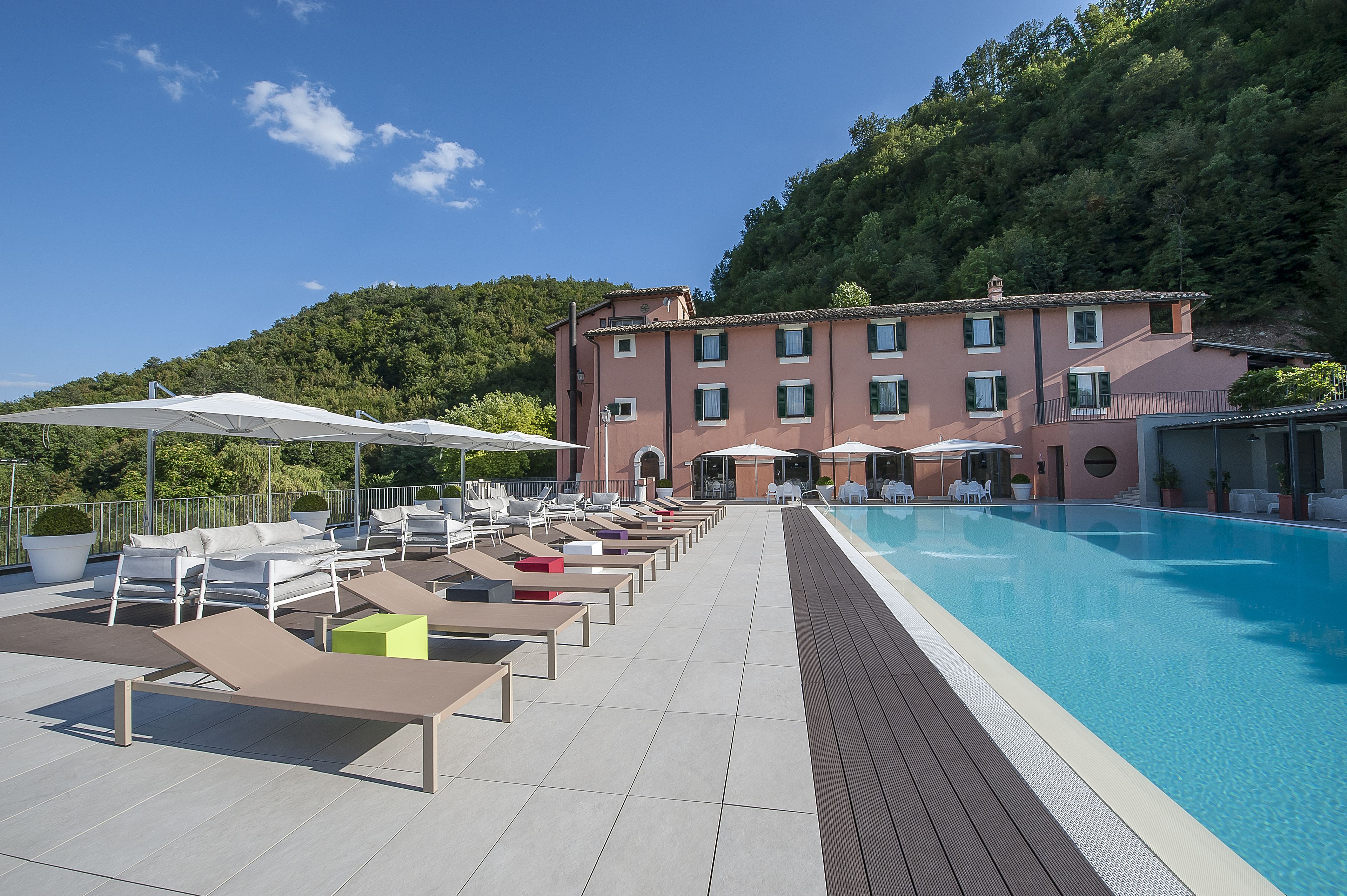 Ein Poolbereich auf dem Dach mit Liegestühlen und Sonnenschirmen, einem wolkenlosen blauen Himmel und einem Gebäude mit Ziegeldach im Hintergrund.