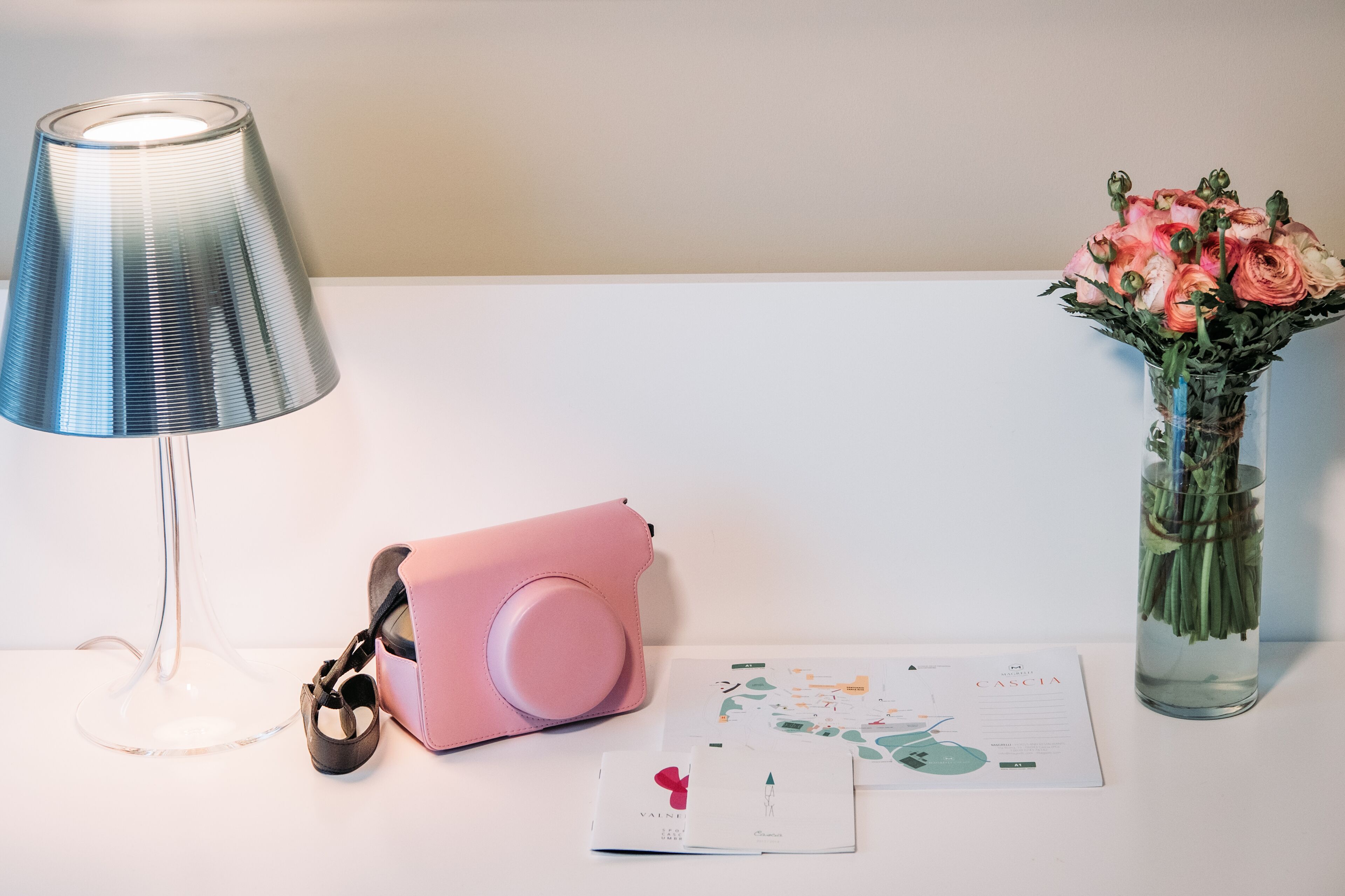 Un tavolo con una lampada, una macchina fotografica rosa, un vaso con fiori e alcuni documenti.