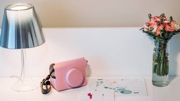 Un tavolo con una lampada, una macchina fotografica rosa, un vaso con fiori e alcuni documenti.
