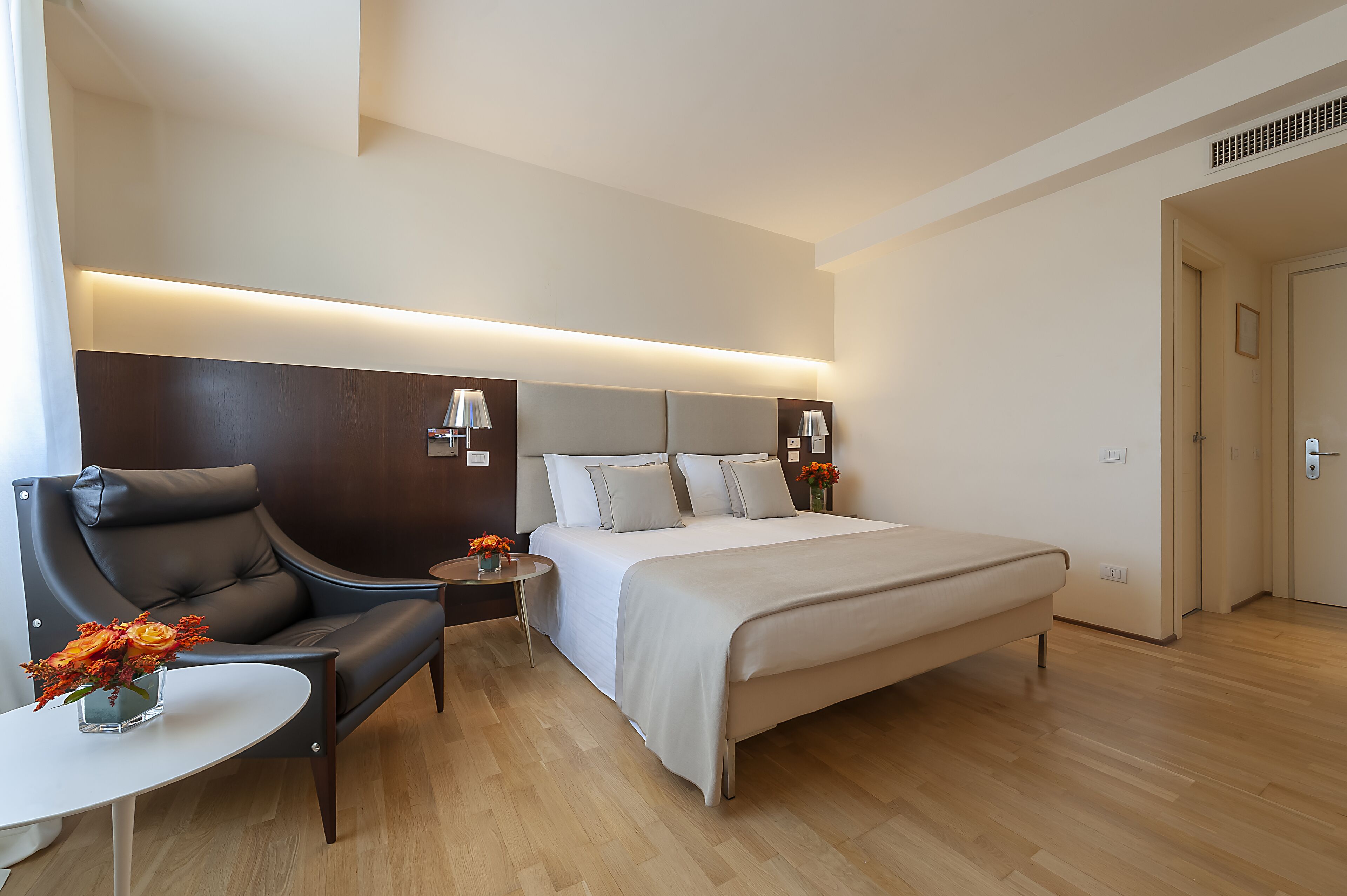 A modern hotel room with a large bed, a leather armchair, a small table, and a bedside lamp.