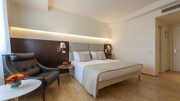 A modern hotel room with a large bed, a leather armchair, a small table, and a bedside lamp.
