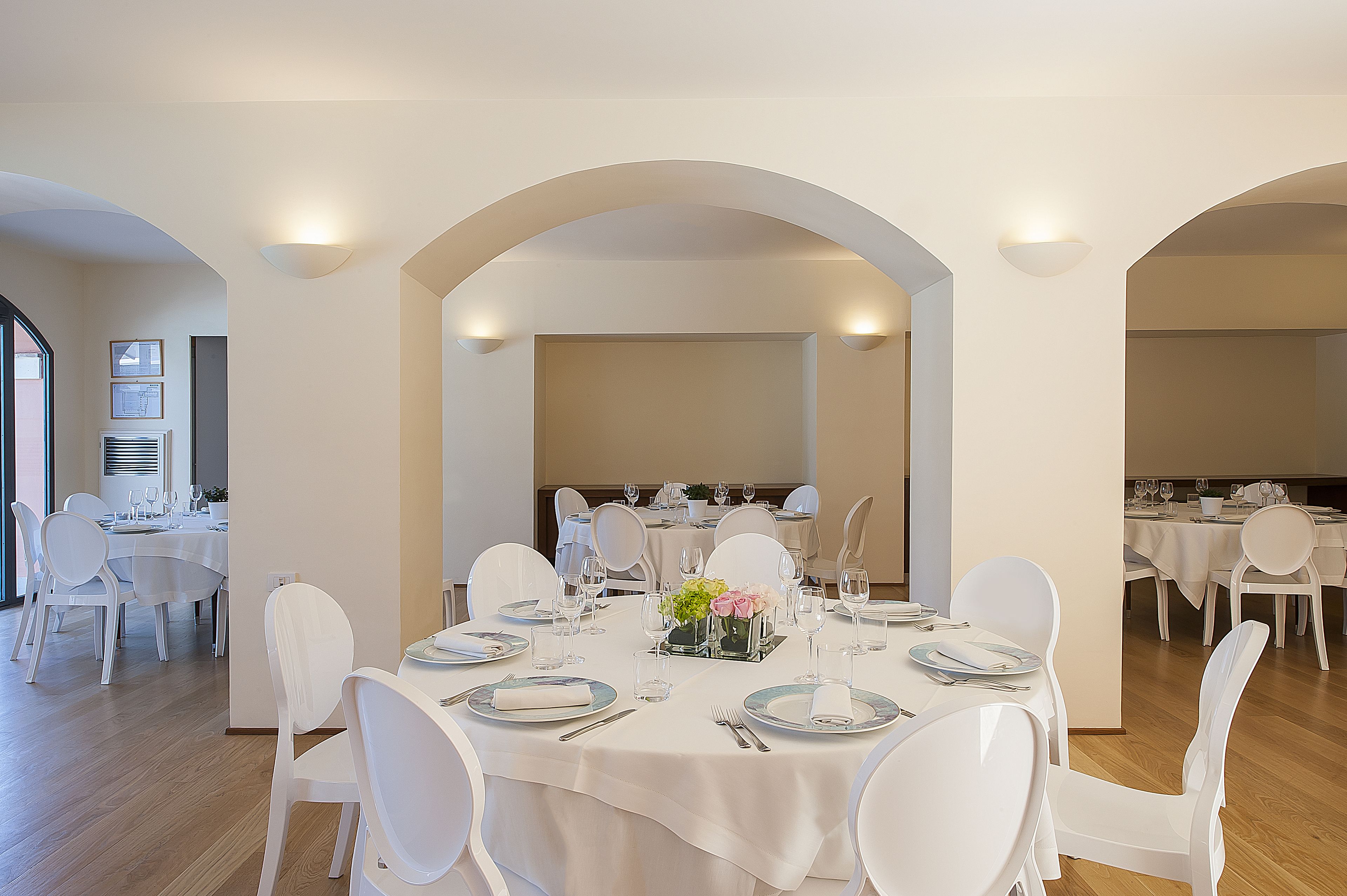 Un comedor con mesas y sillas blancas, dispuesto en una habitación amplia con techos altos y arcos.