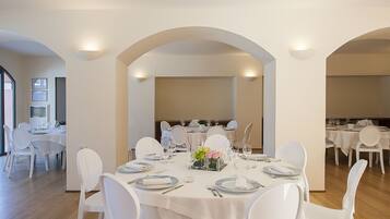 Un comedor con mesas y sillas blancas, dispuesto en una habitación amplia con techos altos y arcos.