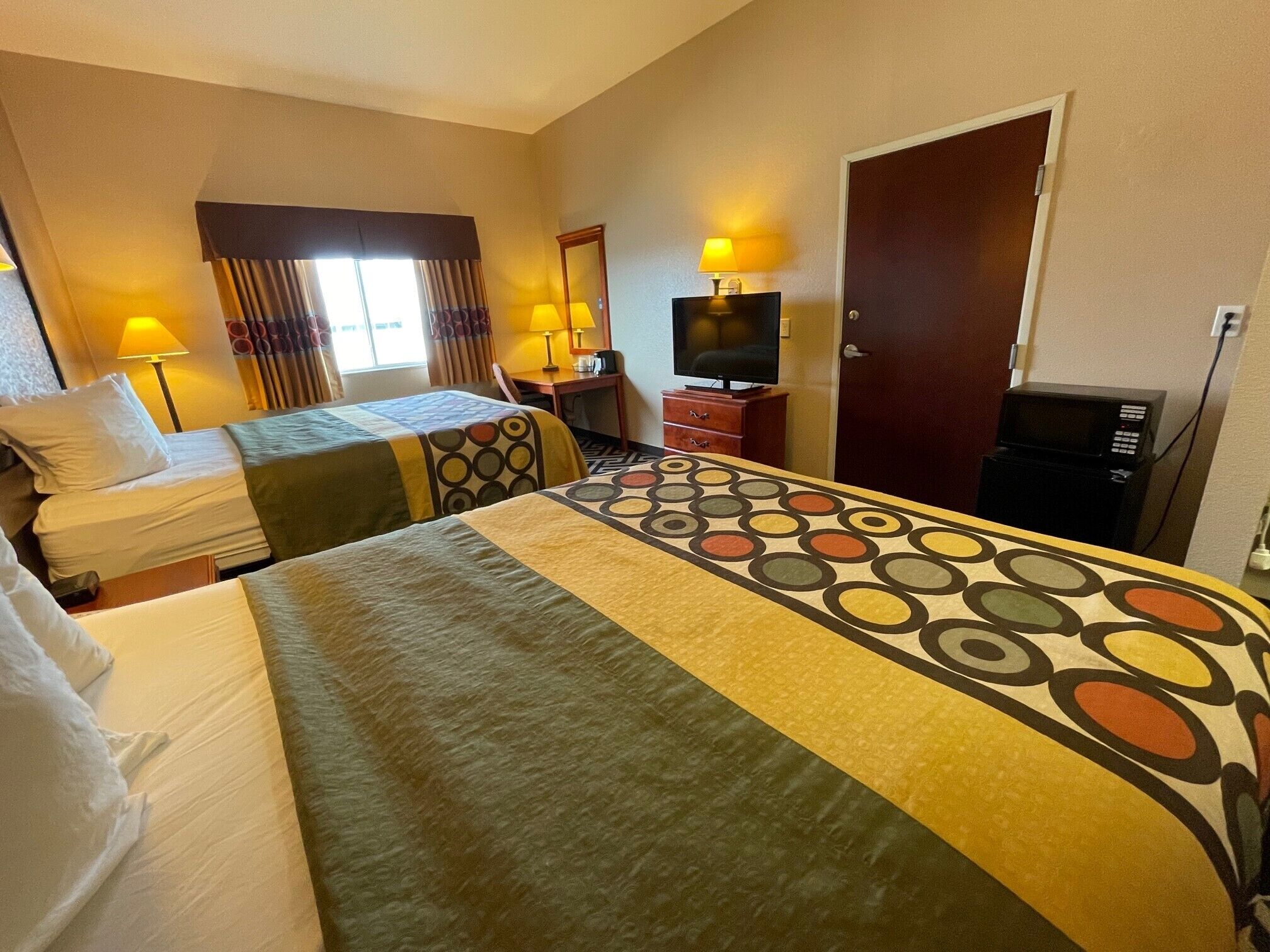A hotel room with two beds, a TV, a desk with a lamp, a door, and a microwave.