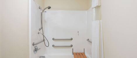 A small, white, walk-in shower with a wooden bench, grab bars, and a showerhead.