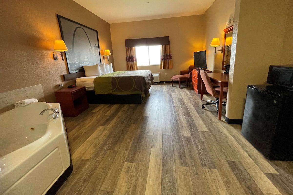 A hotel room with a large bed, a desk, a chair, a desk lamp, a television, a refrigerator, and a bathroom with a bathtub and sink.